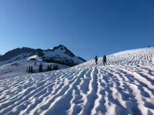 Two hikers walking across a snowy landscape with a mountain peak in the background under a clear blue sky.