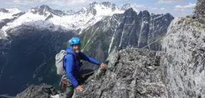 A climber in a blue helmet and jacket ascends a rocky mountain ridge, with snow-capped peaks in the background under a clear sky.