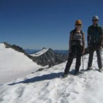 Two climbers wearing helmets and harnesses stand on a snowy mountain ridge, with clear blue skies and distant peaks in the background.