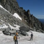 Two hikers with climbing gear on a snowy mountain path, rocky peaks in the background under a clear blue sky.