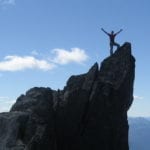 A person stands triumphantly on a mountain peak with arms raised, overlooking expansive mountain views under a clear blue sky.