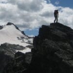 A hiker stands atop a rocky peak, overlooking a snow-covered mountain ridge under a cloudy sky.