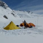 A person standing between brightly colored tents on a snowy mountainous landscape under a clear blue sky.