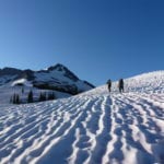 Two hikers walking across a snowy landscape with a mountain peak in the background under a clear blue sky.