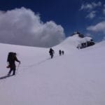 A group of hikers with backpacks ascending a snowy mountain slope under a blue sky with clouds.