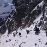 Climbers ascending a snowy mountain slope with rocky cliffs nearby, under a cloudy sky.