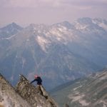 A climber in red helmet ascending a rocky mountain ridge with sweeping views of distant snow-capped peaks and valleys.