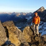 A climber in orange jacket stands on a rocky mountain ridge at sunrise, surrounded by snow-capped peaks under a clear blue sky.