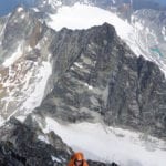 A climber in an orange helmet ascends a rocky mountain ridge with snowy peaks in the background.