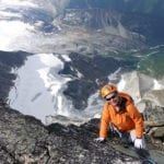 A climber in an orange jacket and helmet ascends a rocky mountain with a glacier visible in the background.