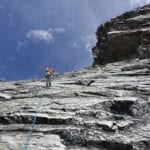 A climber in an orange helmet ascending a steep, rocky mountain face, secured by a blue climbing rope under a clear sky.