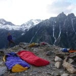 A person standing on a mountain ridge at sunrise, surrounded by sleeping bags and climbing gear, with rugged peaks in the background.