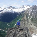 A hiker in a blue jacket stands on a rocky mountain peak, with snow-covered mountains and a deep valley in the background.