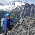 A climber in a blue helmet and jacket ascends a rocky mountain ridge, with snow-capped peaks in the background under a clear sky.