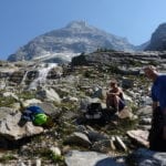 Two hikers taking a break on a rocky mountain trail with a waterfall and a towering mountain in the background.