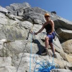 A climber in a helmet and harness stands on a rocky ledge, holding rope, with a steep mountain face in the background.