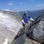 A climber in a blue jacket and helmet scaling a rocky mountain ridge with snow-covered peaks and clear blue sky in the background.