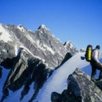 A climber with a backpack stands on a snowy mountain ridge, looking towards sharp, rocky peaks under a clear blue sky.